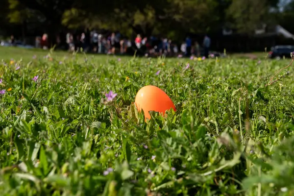An orange, plastic Easter egg hidden in the short grass in a neighborhood park with a grup of people in the blurry background.