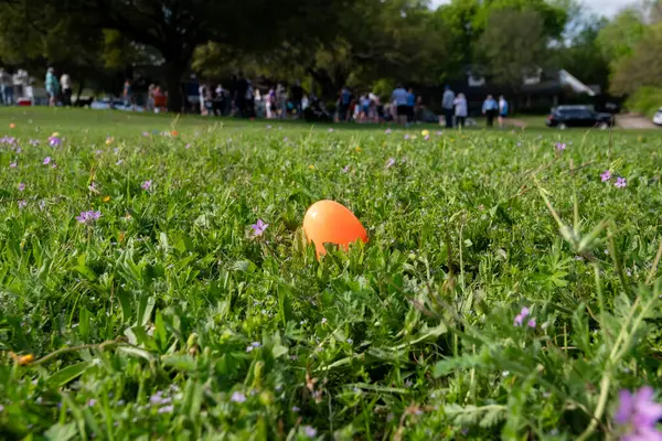 An orange, plastic Easter egg hidden in the short grass in a neighborhood park with a grup of people in the blurry background.