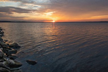 Sıcak turuncu ve sarı bulutlar uzak ufukta güneş batarken gölün sakin suları üzerinde gökyüzünü dolduruyor..