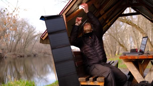 人类在靠近河边的拱门的便携式太阳能电池板上工作 露营用的清洁能源 用便携式太阳能电池板在室外使用小玩意 — 图库视频影像