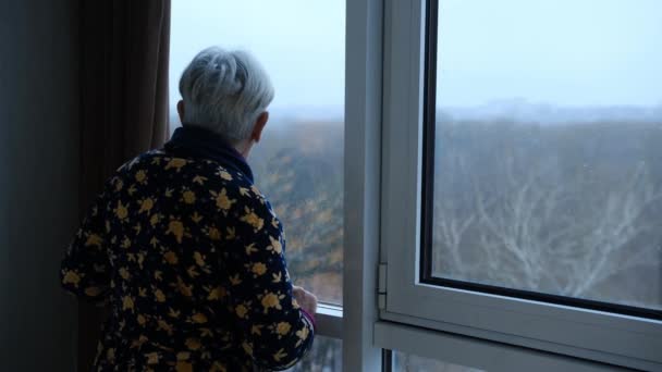 郁闷的老妇人在家里感到难过 老妇人悲伤地看着窗外 孤独的忧郁的女士独自站着 透过窗户看着 — 图库视频影像