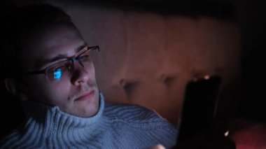 Lonely man in glasses sitting on bed in bedroom and using phone at night. Face illuminated from smartphone display. Surfing social network before sleep.