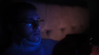 Lonely man in glasses sitting on bed in bedroom and using phone at night. Face illuminated from smartphone display. Surfing social network before sleep.