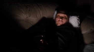 Lonely caucasian middle ages man lie on bed in bedroom and using phone at night. Face illuminated from smartphone display. Surfing social network before sleep.