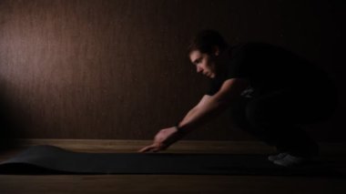 Man in black doing push-ups exercise on the floor on mate at home in a dark room with side lightning.