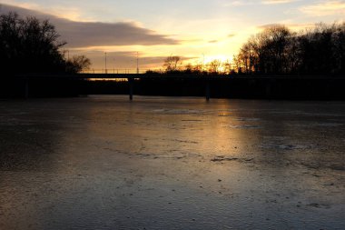 Frosty pink sunset by the frozen river with a bridge going over the water and people walking on it with view from the centre of the frozen river.