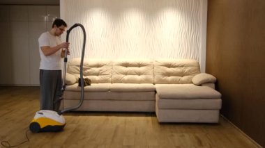 Young caucasian man doing housework at home cat run away when use vacuum cleaner with biege sofa at background.