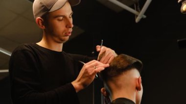 Barber master hairdresser does hairstyle and style with scissors and comb. Concept Barbershop. back view in dark key lightning with warm light. Slow motion