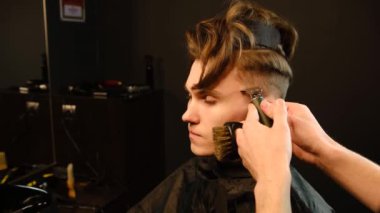 Shot of a handsome barber giving a haircut to his client using trimmer. Hairdresser service in a modern barbershop in a dark key lightning with warm light side view. Slow motion