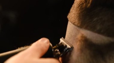 close up Shot of a handsome barber giving a haircut to his client using trimmer. Hairdresser service in a modern barbershop in a dark key lightning with warm light back view.