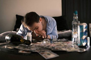 Genç bağımlı keş adam ayna masasında kokain koklarken etrafa saçılmış bir sürü para, alkol, kumar ve haplar yuvarlanmış banknotlar aracılığıyla. Uyuşturucu ve bağımlılık kavramı