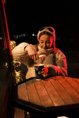 Kırmızı ve sarı bardaklı kahve dükkanının yanında kuyruklu manken sarışın. Kışın kafenin yakınlarında beyaz elbise, ceket ve şapka giymiş bir kız..