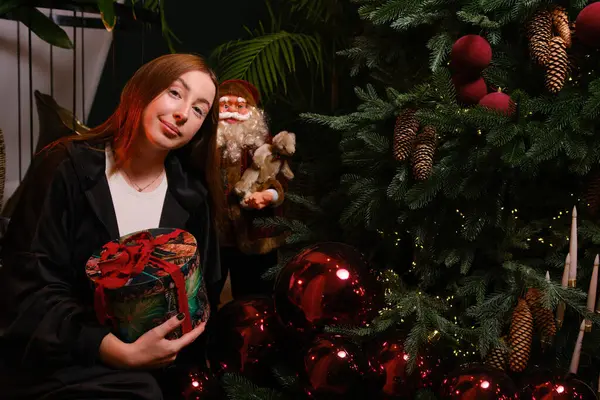 Güzel, genç bir kadın beyaz kirpikleriyle siyah elbiseler içinde Noel ağacının yanında elinde kırmızı şeritli hediyeler ve koyu arkaplanda fiyonk ile oturuyor..