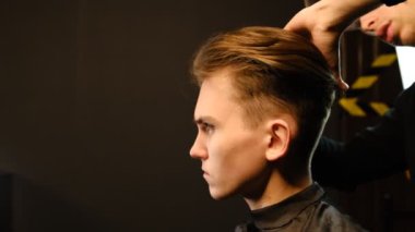 Berber dükkanı. Berber sandalyeli bir adam, kuaför saçını taramak ve şekillendirmek için tarakla hazırlıyor. Berber dükkanı kavramı. Ilık ışıkla karanlık anahtar yıldırımında yan görünüm.