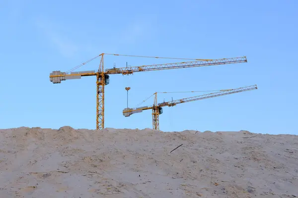 stock image two construction cranes tower over a mound of sand against a backdrop of blue sky. Perfect for illustrating construction progress and industrial development.