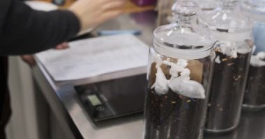 Bir kafe çalışanının çay envanterini yönetirken, şeffaf kavanozlarda çay tartarken, hesap makinesinde toplamları hesaplarken ve detaylara dikkat eden bir formda veri kaydederken yarı bulanık bir kare.