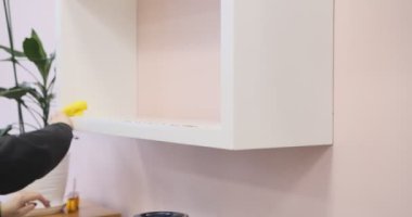 Beyaz bir rafı parlak ve minimalist bir ortamda sarı bir bezle temizleyen kadın elleri pürüzsüz hareketlerle temizliği ve organizasyonu vurguluyor.