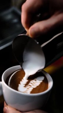 Becerikli barista, beyaz bardakta servis edilen kremalı sütlü sütlü kakao ile rahat bir kafe ortamının arka planında ince bir şekilde görünen profesyonel espresso makinesiyle ayrıntılı bir tekboynuzlu at latte sanatı yaratıyor..