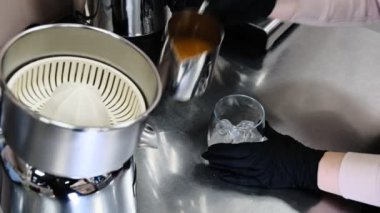 Siyah eldivenli barista, meyve sıkma makinesinin arka planında buzla taze portakal suyunu bardağa boşaltıyor. Meyve sıkma makinesinin yakın çekimi sağlıklı yaşam tarzı C vitaminini tazeliyor.