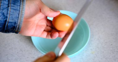 Skillful hands crack an egg with precision using a knife, dropping it into a blue plate set on a sleek grey countertop. Ideal for food preparation tutorials and culinary-themed videos. clipart