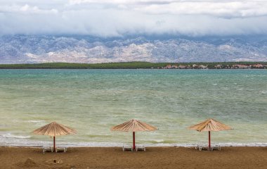 Nin, Hırvatistan - Nin kasabası yakınlarındaki boş Queen 's Beach' te yaz sonunda üç sazlık güneş gözlüğü, arka planda Velebit Dağları ve bulutlu bir sonbahar gününde Adriyatik Denizi turkuaz
