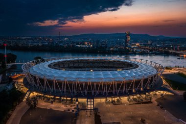 Budapeşte, Macaristan - Tuna Nehri kıyısındaki aydınlık Ulusal Atletizm Merkezi 'nin havadan görünüşü Rakoczi köprüsü, yeni gökdelen binası ve sırt çantasındaki renkli günbatımı gökyüzü