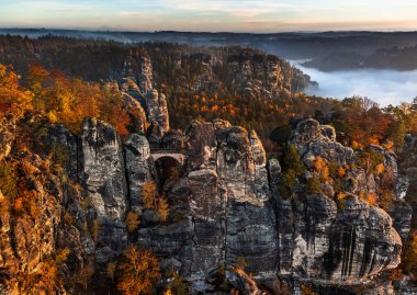 Sakson, Almanya - Sisli bir sonbahar sabahında, kayanın altında yoğun sis ve renkli sonbahar yapraklarıyla Bastei Köprüsü 'nün havadan görünüşü. Bastei, Sakson İsviçre Ulusal Parkı 'nda bir kaya oluşumudur.