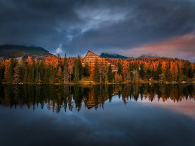 Strbske Gölü, Slovakya - Arka planda Yüksek Tatras ve Tatras Kulesi ile güneşli bir sonbahar öğleden sonrasında ikonik Strbske Gölü 'nü (Strbske Pleso) yansıtan panoramik manzara. Sıcak sonbahar renkleri, bulutlu gökyüzü ve Tatras dağları bulutlarla kaplı.