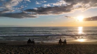 İnsanlar sahilde oturur ve gün batımını izlerler.