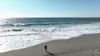 Bir kadın sahilde duruyor ve telefonuna bakıyor. Deniz fırtınası büyük dalgalar gri kum.