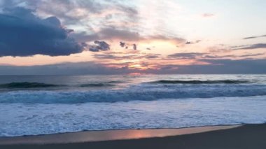 Kumlu sahilde gün batımının resimli görüntüsü. Yumuşak sarı güneş ışığı, yaz akşamı okyanus yüzeyini aydınlatır. Sakin deniz dalgaları boş deniz kıyılarını yıkıyor. Güzel arka plan doğa sükuneti.