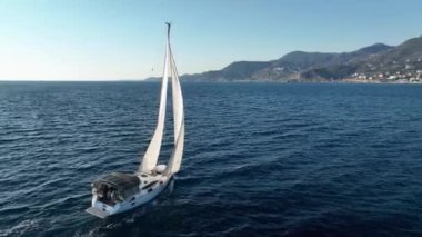 Güneşli bir günde beyaz yelkenli bir yatın sinematik manzarası. Günbatımında Alanya Türkiye kıyılarındaki Akdeniz Koyu 'nda dalgaların üzerinde duran bir yelkenli teknesinin hava görüntüsü.