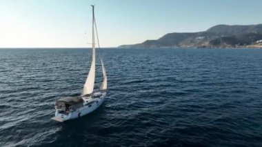 Güneşli bir günde beyaz yelkenli bir yatın sinematik manzarası. Günbatımında Alanya Türkiye kıyılarındaki Akdeniz Koyu 'nda dalgaların üzerinde duran bir yelkenli teknesinin hava görüntüsü.