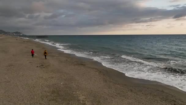 私たちの映画のドローンは ファッショナブルなコートに身を包んだ 日没で絵のような散歩を保存する2人の友人を紹介していますので 冬のビーチの波に身を浸します — ストック動画