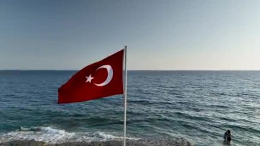 Huzurlu kayalık bir plajın üzerinde rüzgarda zarif bir şekilde dalgalanan Türk bayrağının gururlu görüntüsüne bakın, hayranlık uyandıran bir hava perspektifinden çekildi..