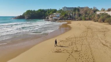 Altın plajda yürüyen bir adamın 4K drone perspektifini izleyin. Akdeniz kıyılarının büyüleyici hava görüntüleri.