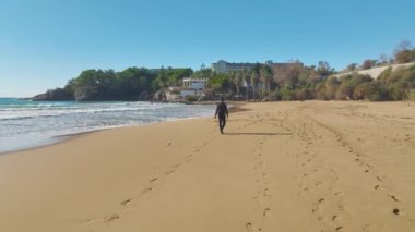 4K hava perspektifiyle genişleyen Akdeniz sahilinde yürüyen siyahlı bir adamın yalnız figürüne bakın..