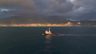 Römorkörün gün batımında limana dönüşünü izleyin, renkli bir gökkuşağı ve gökkuşağı tarafından çerçevelenmiş, çarpıcı bir şekilde 4K.