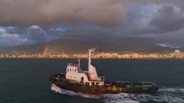 Gün batımında limana dönen bir römorkör görün. Renkli gökyüzü, şehir manzarası ve 4K 'da çarpıcı bir gökkuşağı..