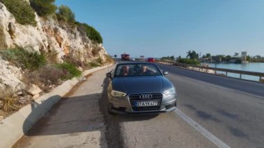 Üstü açık bir arabayla Akdeniz kıyısında, çarpıcı deniz manzaralı bir çift görüyorum..