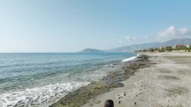 Siyah giyen bir adamın, hava yoluyla 4K 'da yakalanan yumuşak dalgaların arka planında yürürken sakin bir yürüyüş izleyin..