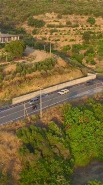 Dikey Video: Bir motosikletçi gün batımı ışığını yansıtan suyun yakınında yolculuk ediyor.