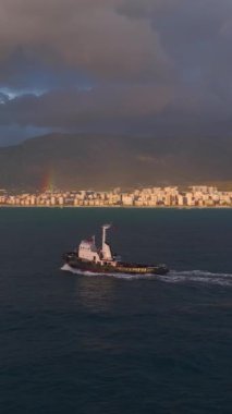 Bir römorkör, gün batımının ve parlak gökkuşağının altında limana doğru yol alır..