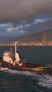 Bir römorkör limana doğru yol alır, alevli bir günbatımı ve parlak gökkuşağı gibi gökyüzünde.