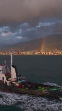 Gökyüzü gün batımı ve gökkuşağı ile aydınlanırken bir römorkör limana doğru yol alır..