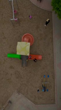 Çocuklar canlı bir oyun bahçesinde oynuyorlar. Yukarıdan görülebiliyorlar. Çevrede yeşillik var..