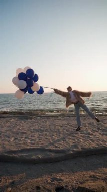 Dikey Video: Moda Model Tutucu Balonlar ve Güneş Ilık Bir Işıltıyla Batarken Deniz kenarında Yürüyüş