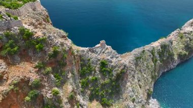 Manzaralı kayalık yarımada denize doğru uzanıyor, korsan teknesi yakınlarda, hava görüntüleri kayalıkları ve sakin suyu yakalıyor.
