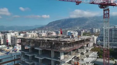 Kıyı inşaatında miğferli işçiler, vinç hareket halindeki levhalar, arka planda Akdeniz ve şehir.