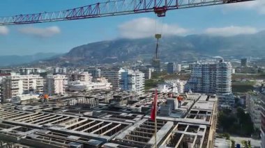 Drone görünümü: vinç malzemeleri indiriyor, çerçeve inşa eden işçiler, deniz kenarındaki ufuk çizgisi geriden açılıyor.